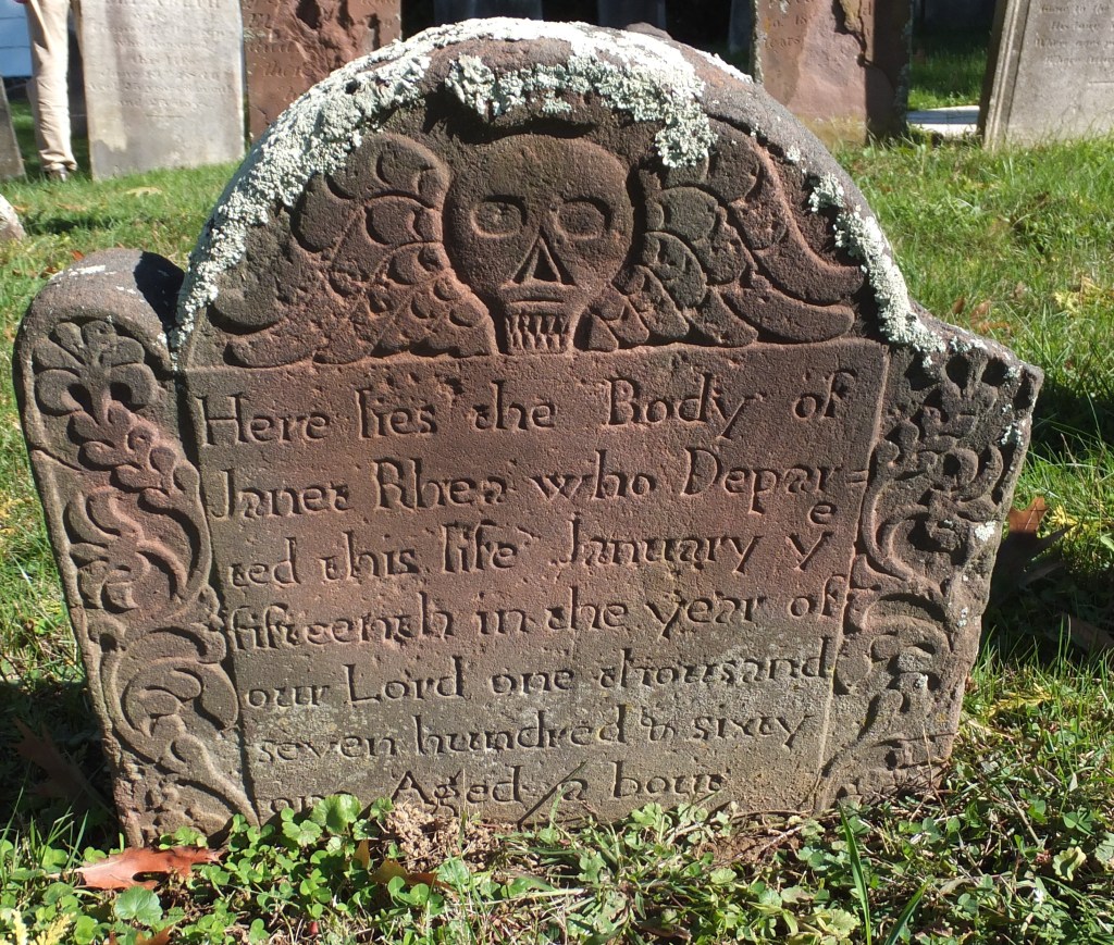 An old stone gravestone with a skull motif and intricate floral designs, inscribed with the epitaph of Janet Rhea, who passed away on January 15, 1760, aged 62.